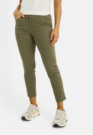 Persona con pantalones ajustados cortos de verde oliva y zapatillas beige-blancas, de pie con una mano en el bolsillo contra un fondo blanco.