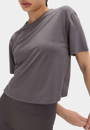 Kvinde iført en løs, kortærmet, grå crop-top og tætsiddende mørkegrå leggings mod en ensfarvet hvid baggrund.