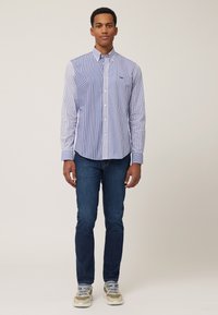 Blau-weiß gestreifte Hemdbluse mit einer einzelnen Brusttasche, kombiniert mit einer dunklen Jeans und beigen Sneakers mit blauen Akzenten.