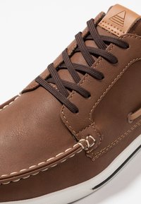 Chaussure décontractée en cuir marron avec des lacets brun foncé, des surpiqûres blanches le long de l'avant-pied et une semelle en caoutchouc blanche avec une bande noire.