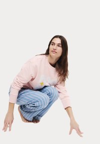 Ljusrosa sweatshirt med vit text, tillsammans med blåa och rosa randiga loungebyxor, visad i en hukande position.