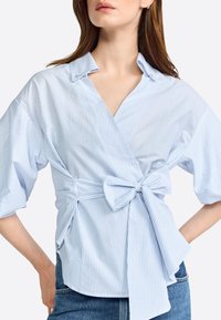 Hellblau gestreiftes Wickelshirt mit einer Schleifenakzentuation an der Taille, kurzen Ärmeln und Kragen. Der Stoff wirkt glatt mit vertikalen Streifen.