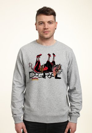 Mand iført en grå sweatshirt med en tegneseriefigur Deadpool, der ligger på hovedet, og teksten "Deadpool Wolverine" på fronten.