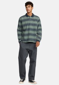 Langarm-Polohemd in gedämpften Grüntönen mit Blau gestreift, ausgestattet mit einem Kragen, einem Emblem auf der Brust und lockerem Schnitt. Kombiniert mit lockeren grauen Hosen und Turnschuhen.