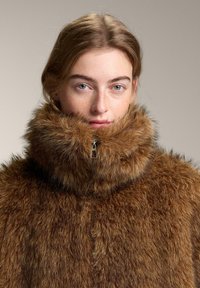 Műszőrméből készült barna kabát magas gallérral és cipzárral. Puha, texturált felülettel rendelkezik, vastag, bolyhos megjelenéssel.