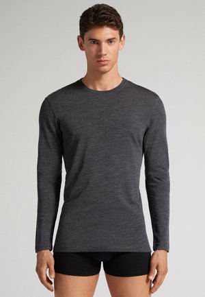 Junger Mann mit kurzen Haaren, der ein enganliegendes dunkelgraues Langarmshirt und schwarze Shorts trägt, steht vor einem schlichten hellen Hintergrund.