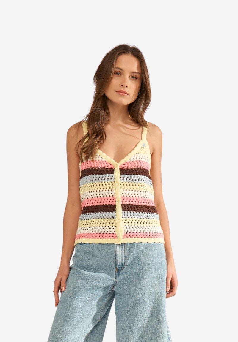 Femme portant un débardeur en crochet multicolore avec boutons, associé à un jean en denim bleu clair ample, debout devant un fond blanc.