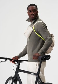 Khaki Zip-Cardigan, creme Umhängetasche mit einem leuchtend grünen Gurt und beigem Rucksack. Schwarzes Fahrrad mit schickem Design.