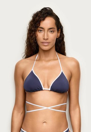 Kobieta o kręconych włosach, ubrana w granatowy top od bikini z białym obramowaniem i skrzyżowanymi ramiączkami na jednolitym jasnym tle.