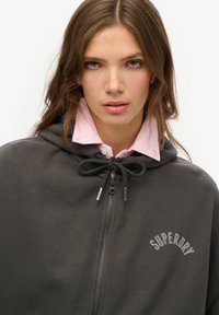 Dunkelgrauer Kapuzenumhang mit zip-front, Zugkordelverschluss und "SUPERDRY"-Druck in hellerem Grau. Weiche Textur, über einem rosa Hemd mit Kragen getragen.