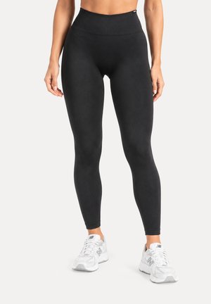 Femme portant un legging noir taille haute et des baskets blanches New Balance, debout devant un fond clair uni.