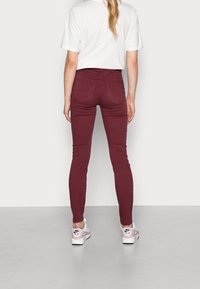 Jean skinny bordeaux, en tissu extensible. Poches arrière visibles. Associé à une chemise blanche et des baskets claires. Texture lisse.