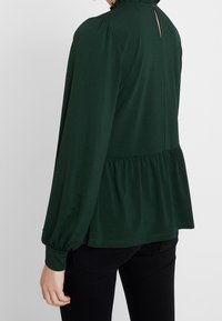 Tmavě zelená blůza s texturovaným povrchem, vysokým límcem a nařaseným peplum lemem. Dlouhé rukávy s elastickými manžetami. Otevření ve tvaru klíčové dírky na zadní straně.