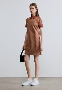 Rövid ujjú, rozsdabarna pólóruha ismétlődő geometriai mintával, piros gallérral és oldalsó csíkokkal. Fehér sportcipővel és fekete táskával viselve.