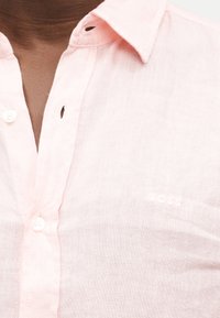 Κοντινό πλάνο ενός ατόμου που φοράει ανοιχτό ροζ πουκάμισο με γιακά και κουμπιά, με τρία κουμπιά εμφανή και διακριτική κεντητή στάμπα "BOSS".