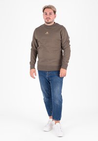 Oliwkowy sweter z okrągłym dekoltem i ściągaczami na rękawach, zestawiony z jasnoniebieskimi dżinsami i białymi trampkami. Złoty akcent w postaci logo na klatce piersiowej.