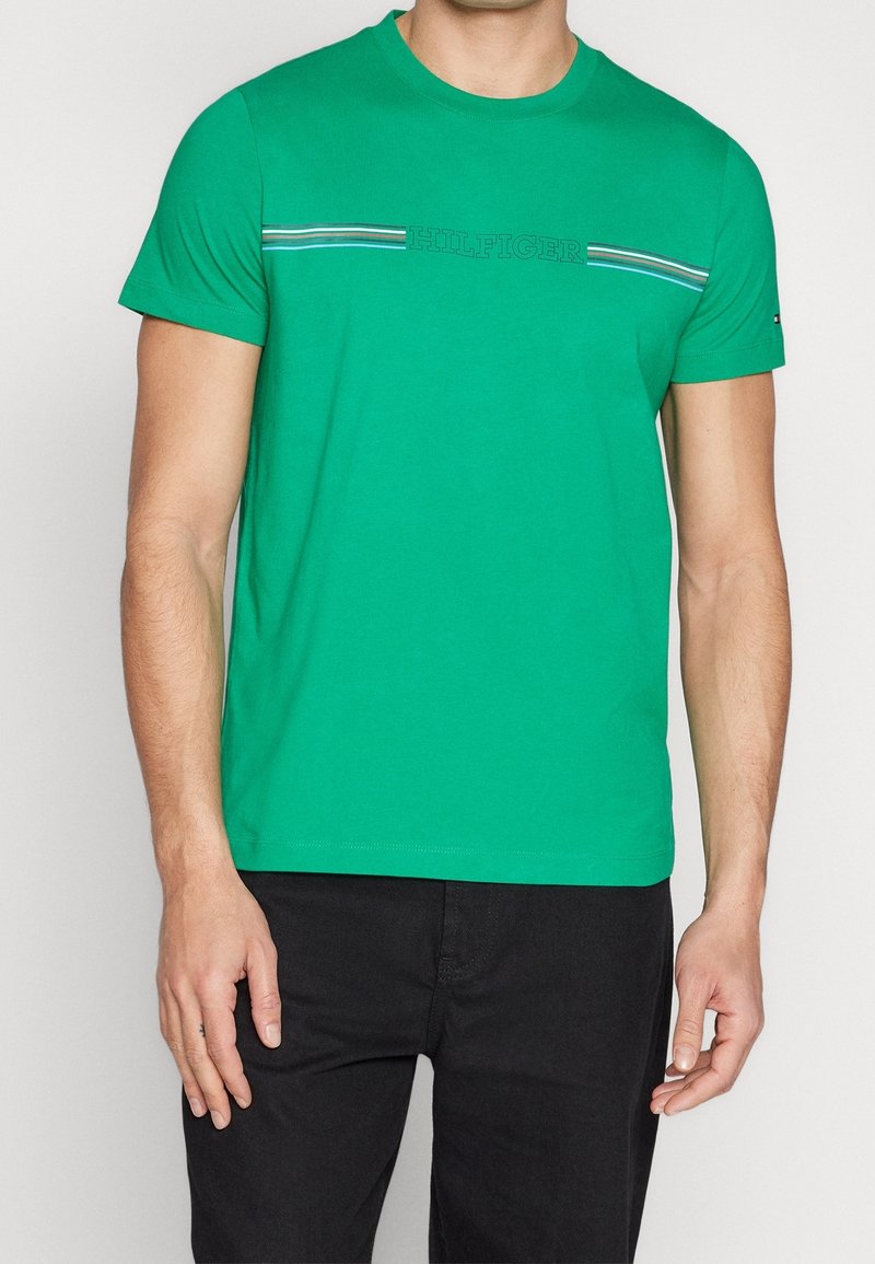 Man wearing a green short-sleeve t-shirt with "HILFIGER" text and multicolored stripes across the chest, paired with black pants.