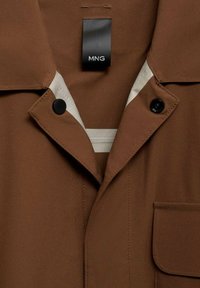 Brown lightweight jacket with a classic collar, black buttons, a chest pocket, and a contrasting white interior lining.