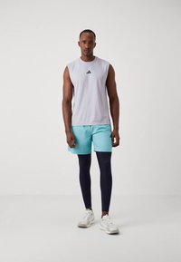 Mouwloze grijze sporttop met zwart logo, gecombineerd met aqua kleurige shorts en marineblauwe legging. Witte sneakers maken de outfit compleet.