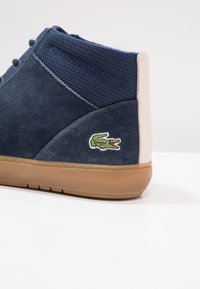 Basket en daim bleu marine et mesh avec languette beige au talon, semelle en caoutchouc et logo crocodile vert sur le côté.