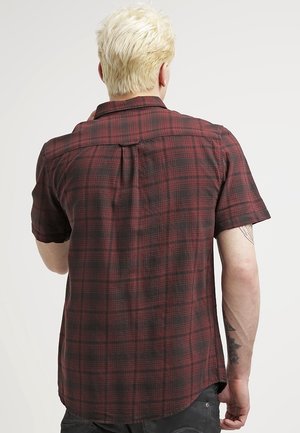 Blond person viewed from behind wearing a short-sleeve red and black plaid shirt, showing tattoos on the right forearm.