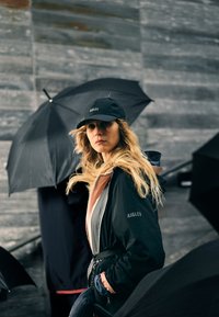Giacca impermeabile nera con chiusura a zip, indossata sopra un top rosa; cappellino da baseball nero. Circondata da ombrelli neri su uno sfondo grigio.