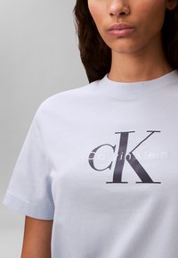 Vrouw draagt een lichtblauw T-shirt met het zwart-witte Calvin Klein-logo in het midden van de borst, tegen een effen grijze achtergrond.