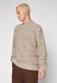 Šviesiai smėlinis megztinis su tekstūruotu deimantų raštu, apvaliu iškirpimu, įlinkusių rankovių ir liemens juosta. Dėvimi su rudais cargo džinsais.