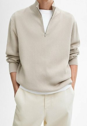 Mann trägt einen beigen, gerippten Pullover mit Viertel-Reißverschluss und einem weißen T-Shirt darunter, die Hände in den Taschen seiner cremefarbenen Hose.