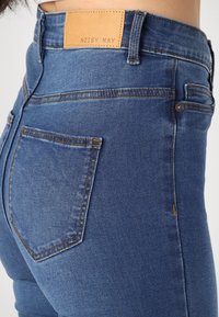 Close-up de uma pessoa a usar jeans de cintura alta azuis, com costuras visíveis, bolso traseiro e uma etiqueta de marca de couro castanho "Noisy May."