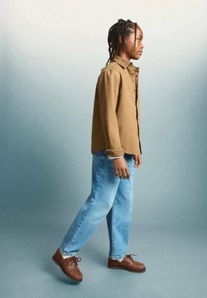 Bambino con capelli intrecciati che cammina di lato indossando una camicia beige con bottoni, jeans azzurri e scarpe marroni con lacci su uno sfondo grigio uniforme.