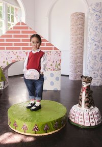 Litet barn med glasögon, röd väst och jeans står på en grön cirkulär plattform med en nallebjörnsskulptur i närheten i ett dekorerat inomhusrum.