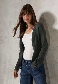 Dunkelgrüne, gerippte Strickjacke mit Knopfverschluss, getragen über einem weißen Oberteil. Kombiniert mit blauen Jeans. Die Textur ist gestrickt; lässige Passform.