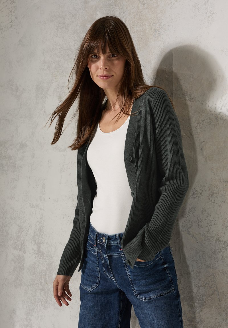 Dunkelgrüne, gerippte Strickjacke mit Knopfverschluss, getragen über einem weißen Oberteil. Kombiniert mit blauen Jeans. Die Textur ist gestrickt; lässige Passform.
