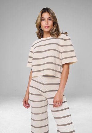 Donna con capelli alla lunghezza delle spalle che indossa una maglia a maniche corte beige e taupe a righe orizzontali e pantaloni larghi abbinati, in piedi davanti a uno sfondo grigio.