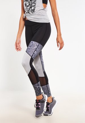 Leggings taille haute en noir, gris et imprimé serpent. Comprend des panneaux en maille et un design ajusté. Portés avec des chaussures de sport grises.
