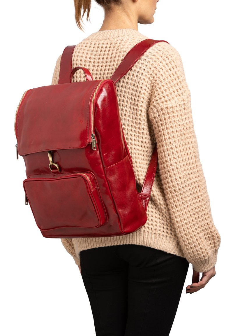 Vrouw draagt een beige gebreide trui en zwarte broek en heeft een grote glanzende rode leren rugzak met voorvak en klepsluiting bij zich.