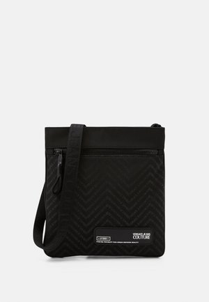 Schwarze Umhängetasche mit vorderem Reißverschluss, verstellbarem Riemen, Chevron-Muster und Versace Jeans Couture Logo-Patch.