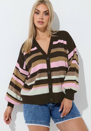 Vrouw draagt een bruine, roze, beige en witte gestreepte, geknoopte cardigan met ballonmouwen en blauwe denimshorts met kanten rand.