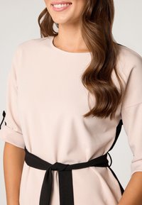 Różowa sukienka o okrągłym dekolcie, z rękawami o długości 3/4, z czarnym materiałowym paskiem związanym w talii, charakteryzująca się gładką fakturą.