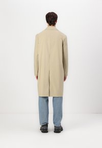 Beige lang frakke med lige snit, enkelt slidssprække bagpå, parret med lyseblå jeans og sorte sko, set bagfra.