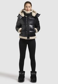 Zwarte pufferjack met een zachte crèmekleurige binnenvoering, hoge kraag en oversized capuchon. Gecombineerd met zwarte leggings en zwarte winterlaarzen.