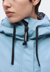 Gros plan d'une personne portant une veste à capuche bleu clair zippée avec des cordons noirs et un curseur de fermeture éclair de marque.