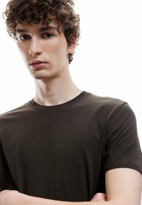 Ciemnobrązowa bawełniana koszulka typu crew-neck z krótkimi rękawami, gładka w dotyku, z subtelnymi detalami przeszycia. Model pokazany od ramion w górę.