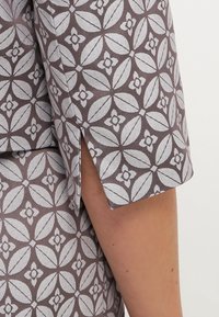 Koszulka z krótkim rękawem w brązowo-białym geometrycznym wzorze kwiatowym, z małym rozcięciem po bokach powyżej łokcia, odsłaniającym nagą rękę.