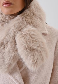 Manteau en laine rose clair avec un col en fausse fourrure doux dans une teinte similaire, présentant une finition texturée et de grands détails duveteux.