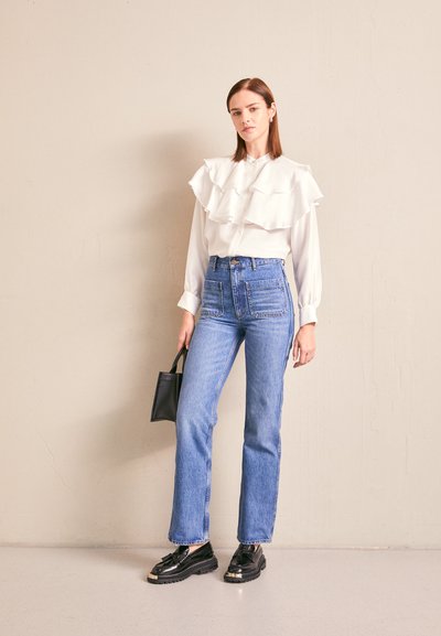 Mujer vestida con una blusa blanca con volantes, jeans azules de cintura alta, mocasines negros y sosteniendo un pequeño bolso negro contra una pared lisa.