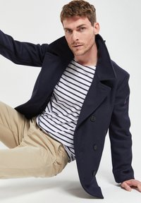 Cappotto peacoat in lana blu navy con ampio colletto e chiusura doppiopetto, indossato sopra una camicia a righe. I chinos beige completano l'outfit.