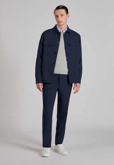 Chaqueta azul marino con botones y dos bolsillos delanteros, usada sobre un suéter gris, combinada con pantalones azul marino a juego y zapatillas blancas.