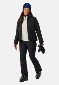 Sort isoleret jakke med hætte, beige zip-up trøje, sorte skibukser og sorte skistøvler. Skibriller bæres på hovedet.